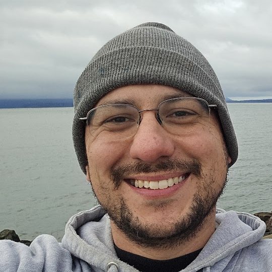 Chris Mendoza, wearing a gray beanie and hoodie, on the Puget Sound shoreline.
